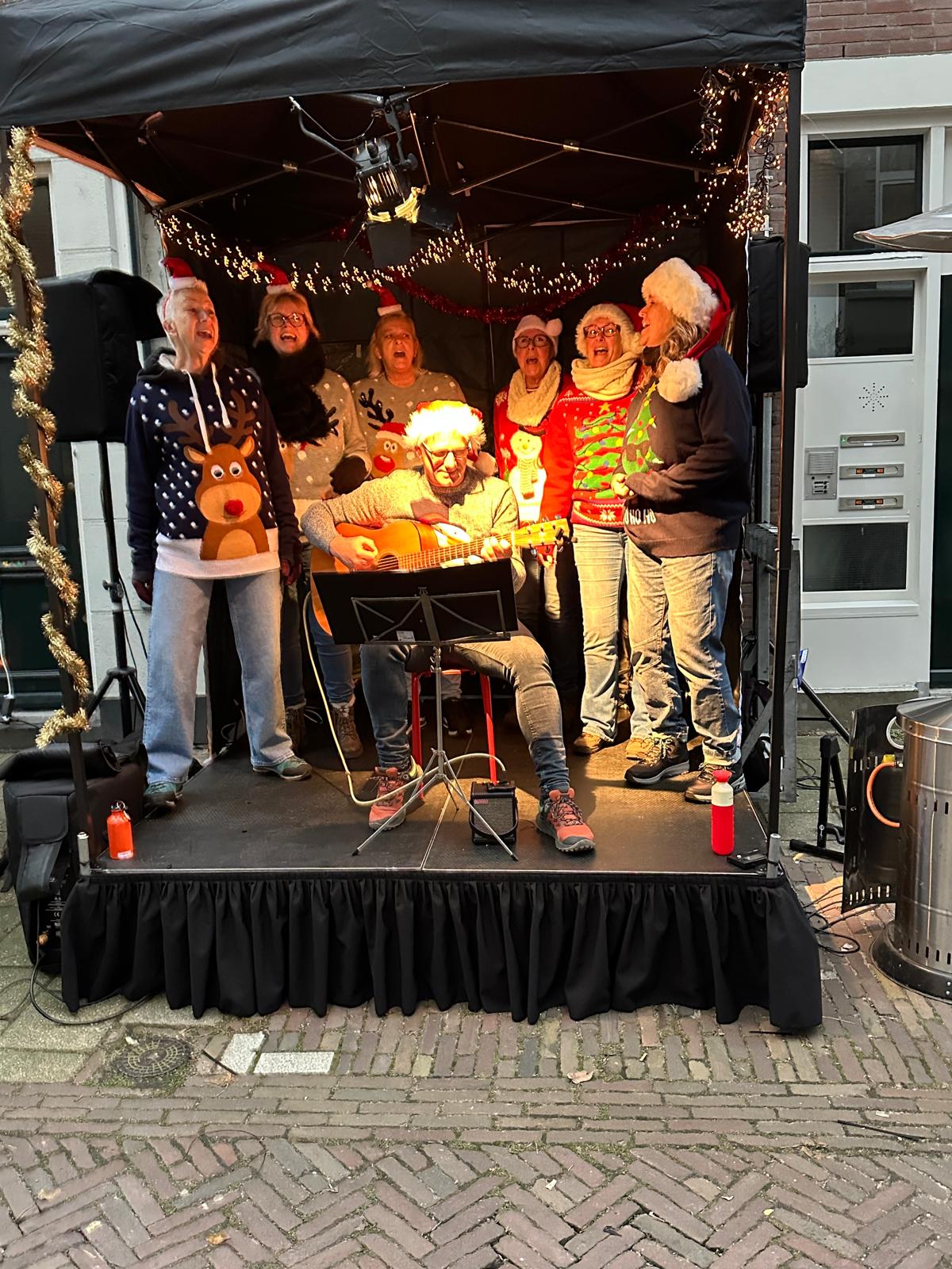 25 koorleden staan lachend achter een grote afbeelding van rode ouderwtse amerikaanse auto.